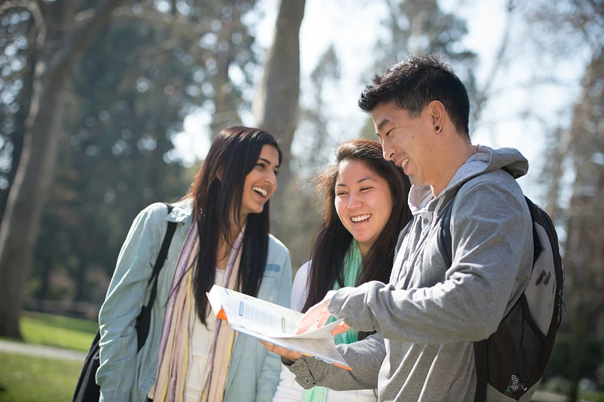 Three college students looking at a book, outside on campus